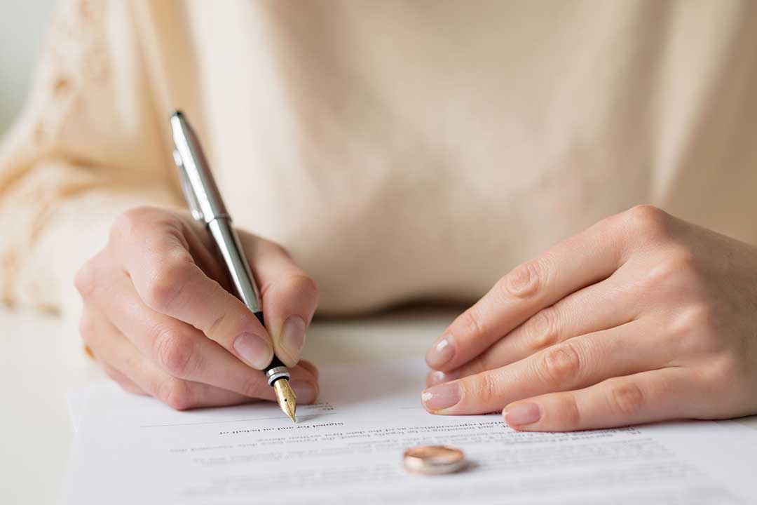 Person signing a legal agreement on a document with a wedding ring placed nearby